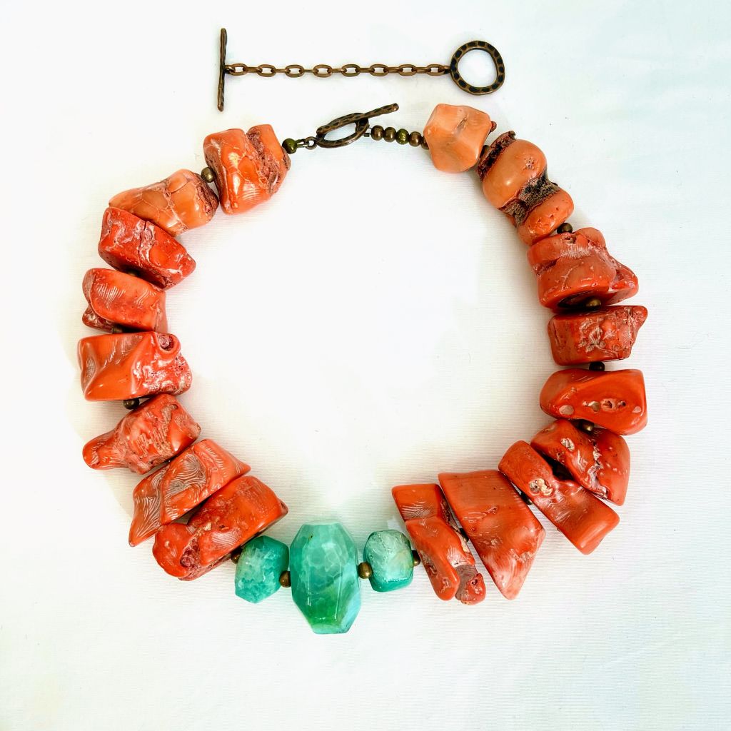 Handcrafted Chunky Coral Necklace displayed on a white background, featuring large irregular orange coral beads with natural texture and three faceted turquoise‑green agate stones at the center. The necklace includes a bronze toggle clasp, highlighting its artisanal craftsmanship and bold color contrast.