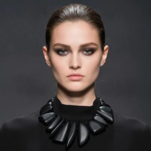 Model wearing a sleek black high‑neck top and a bold sculptural black statement necklace made of smooth, asymmetrical pieces. The minimalist dark background highlights the necklace’s glossy texture and modern design.