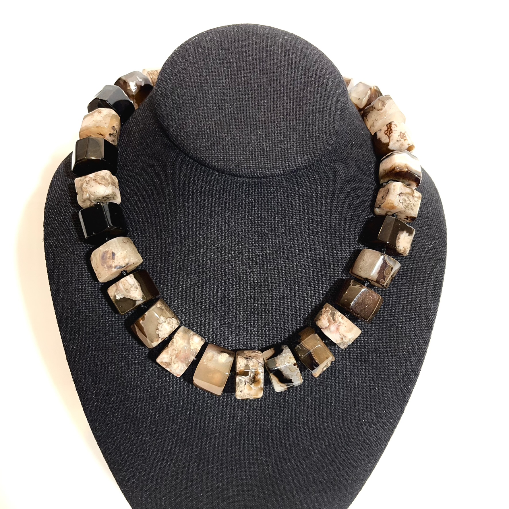 Handcrafted necklace featuring alternating faceted brown and cream agate stones with natural marbling patterns, displayed on a black jewelry bust. The design highlights earthy tones and geometric craftsmanship.