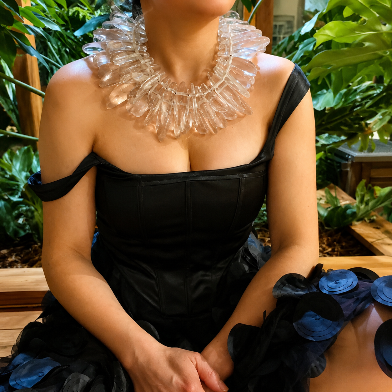 Model wearing a handcrafted Clear Sculptural Bead Necklace featuring elongated transparent acrylic beads arranged in a dramatic circular design. Styled with an off‑shoulder black dress in a lush indoor setting, showcasing the necklace’s bold, artistic elegance and luminous texture.