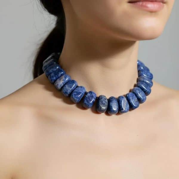 Blue Necklace Bold, beautiful, and effortlessly chic—the Handcrafted Blue Necklace of Sodalite is a wearable celebration of color, craftsmanship, and confidence. Featuring large, irregular-shaped semiprecious Sodalite stones, this standout accessory adds a pop of rich royal blue to any ensemble. Each natural stone is individually knotted by hand, ensuring both durability and comfort while creating a distinctive drape that lies gracefully along the neckline. At first glance, its vivid hue evokes the deep elegance of Lapis Lazuli. Yet, Sodalite offers its own unique charm—its royal blue tone is softer, more grounded, and free from the golden flecks of pyrite, making it a subtler, more versatile choice for day-to-night wear. The natural variations in the stones’ shape and texture highlight the necklace’s artisanal character, giving each piece its own personality and visual rhythm. Perfect for the fashion-forward woman who embraces bold details with refined taste, this blue necklace effortlessly enhances everything from structured blazers to flowing dresses. It’s more than just an accessory—it’s a statement of individuality, a touch of handcrafted artistry, and a stylish nod to the timeless allure of blue stones. Versatile, eye-catching, and utterly wearable, the Blue Necklace of Sodalite is poised to become your new favorite go-to piece.