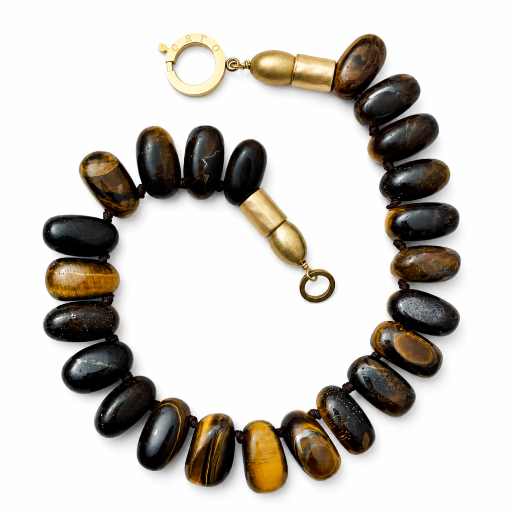 Handcrafted necklace featuring large polished Tiger Eye beads in golden‑brown tones with chatoyant bands, accented by matte gold cylindrical beads and a circular clasp engraved “Caro.” Displayed on a light background.