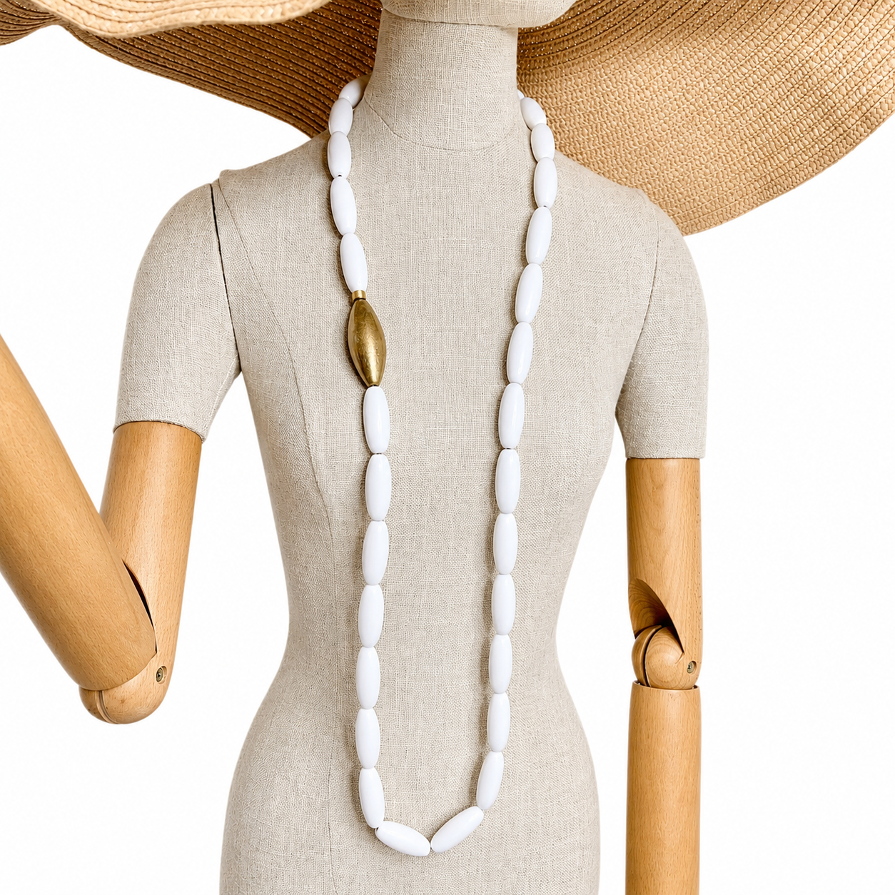 Mannequin wearing a long white bead necklace with a single gold accent bead, styled with a wide‑brimmed straw hat for a minimalist Bohemian look.