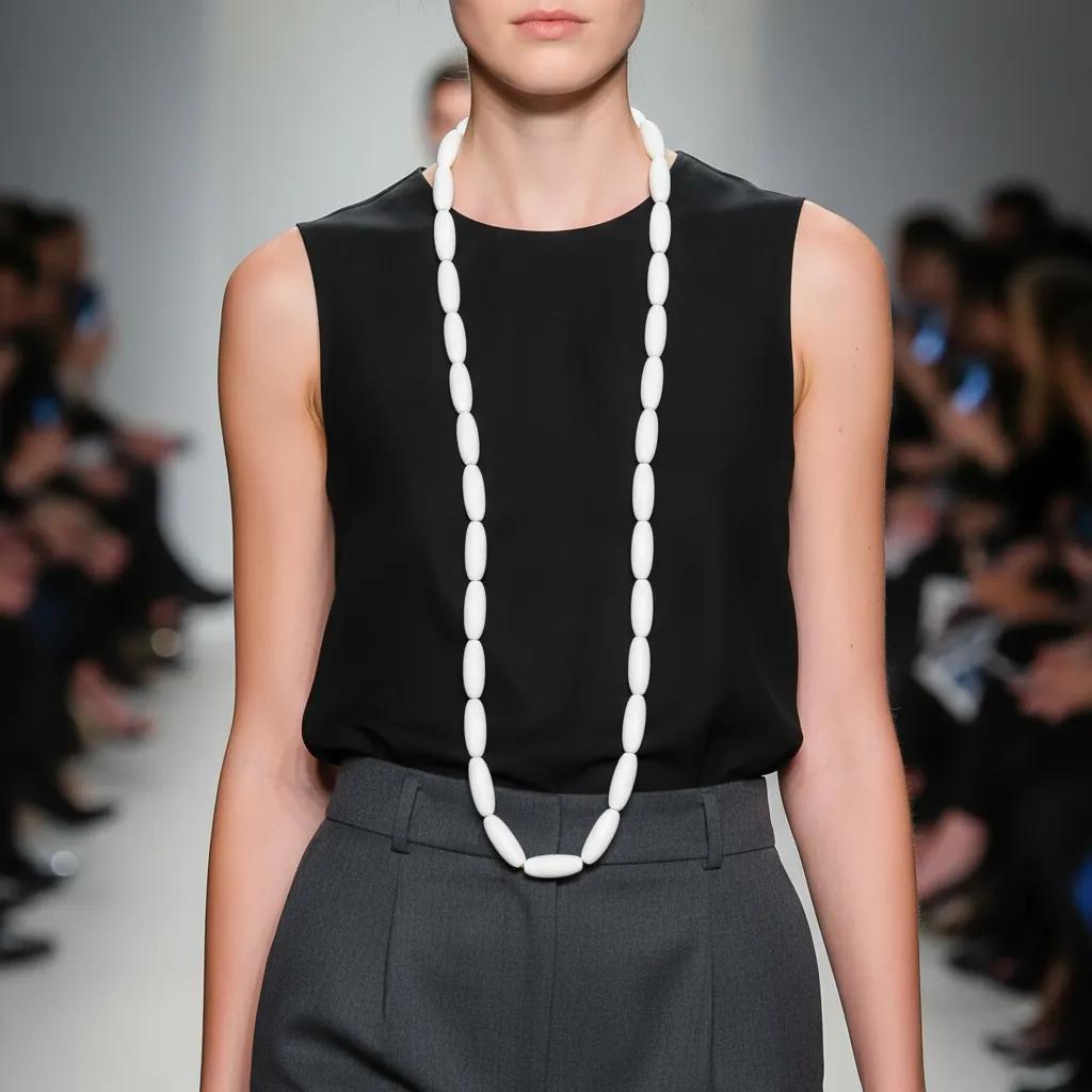 Model wearing a minimalist long white bead necklace over a sleeveless black top and gray trousers on a runway, showcasing elegant contrast and modern style.