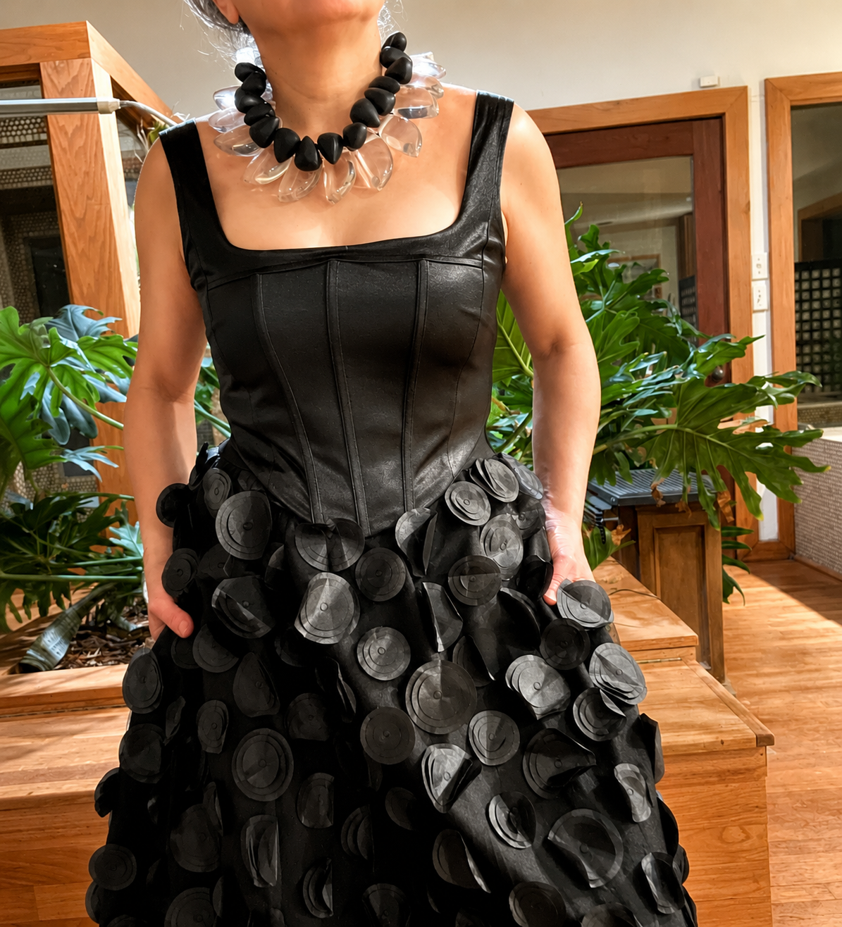 Model wearing a bold collar necklace with black and clear beads, styled with a black leather corset dress featuring circular appliqués in an elegant indoor setting.