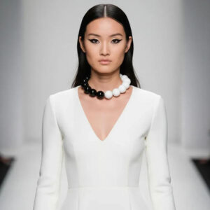 Model wearing a black and white bead necklace with a minimalist white V‑neck dress on a runway, showcasing monochrome elegance and bold contrast.