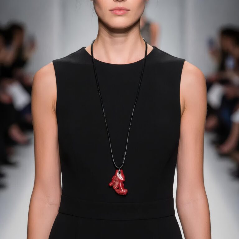 Model wearing a long black cord necklace with a vivid red coral pendant over a sleeveless black dress on a runway.