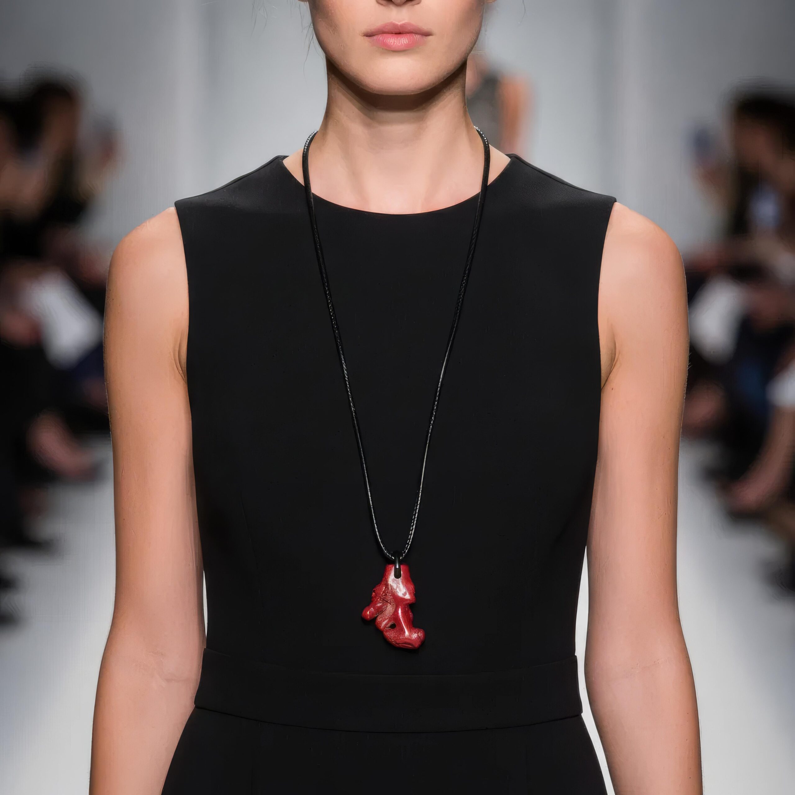 Model wearing a long black cord necklace with a vivid red coral pendant over a sleeveless black dress on a runway.