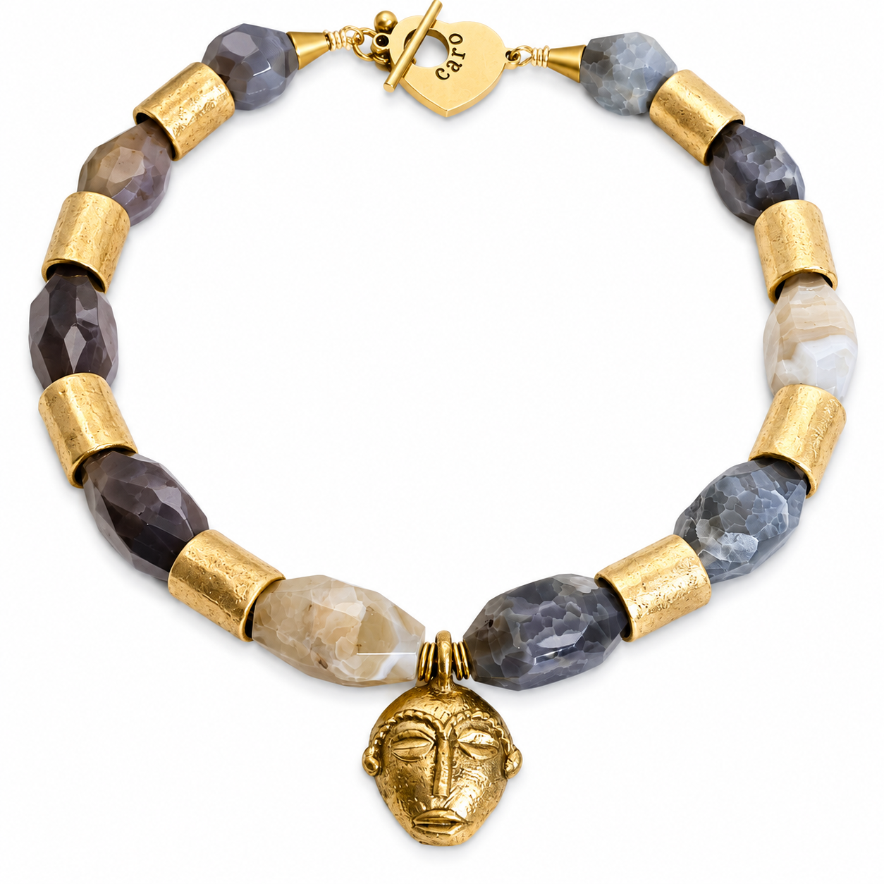 Handcrafted necklace featuring faceted gray and beige crystal beads interspersed with cylindrical gold metal spacers, centered by a sculpted gold face pendant, displayed on a white background with a gold toggle clasp engraved “Caro.”