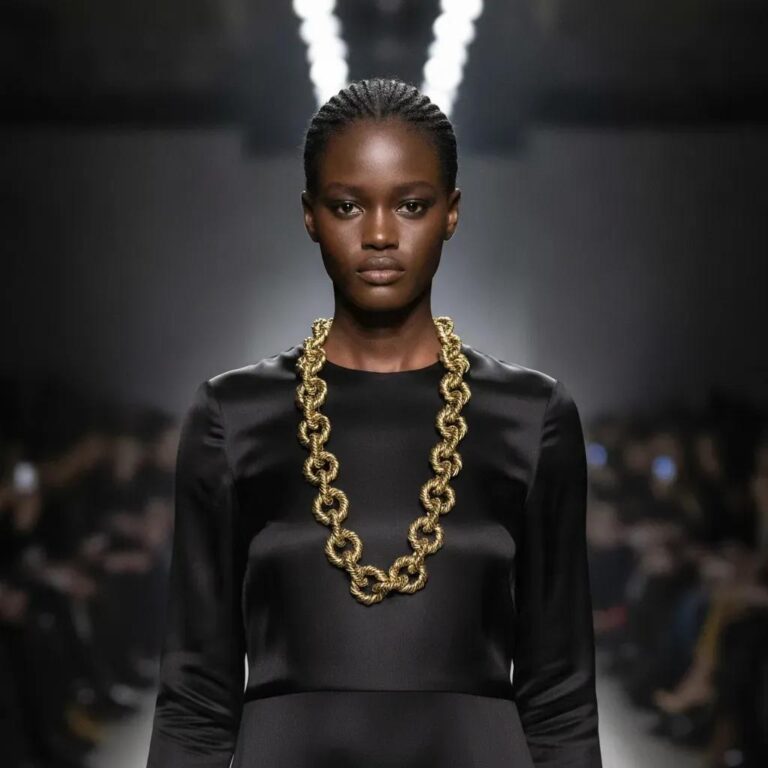 Model wearing a long gold rope‑link chain necklace with large twisted circular links over a black satin dress on runway.