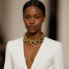 Model wearing a bold gold rope‑link necklace with large twisted circular links, styled with a white blazer and sleek hair.