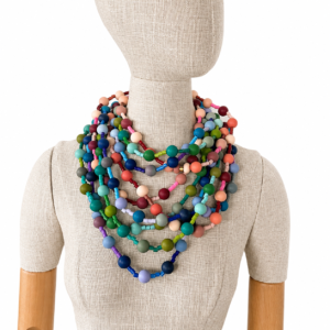 Mannequin wearing multiple layered long necklaces made of round and cylindrical silicone beads in a wide range of colors—blue, green, pink, peach, purple, and beige—creating a vibrant, stacked look against a neutral fabric background.
