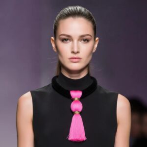 Model wearing a striking necklace with a thick black suede collar and a large hot pink silk tassel pendant, styled over a sleeveless black dress on a runway.