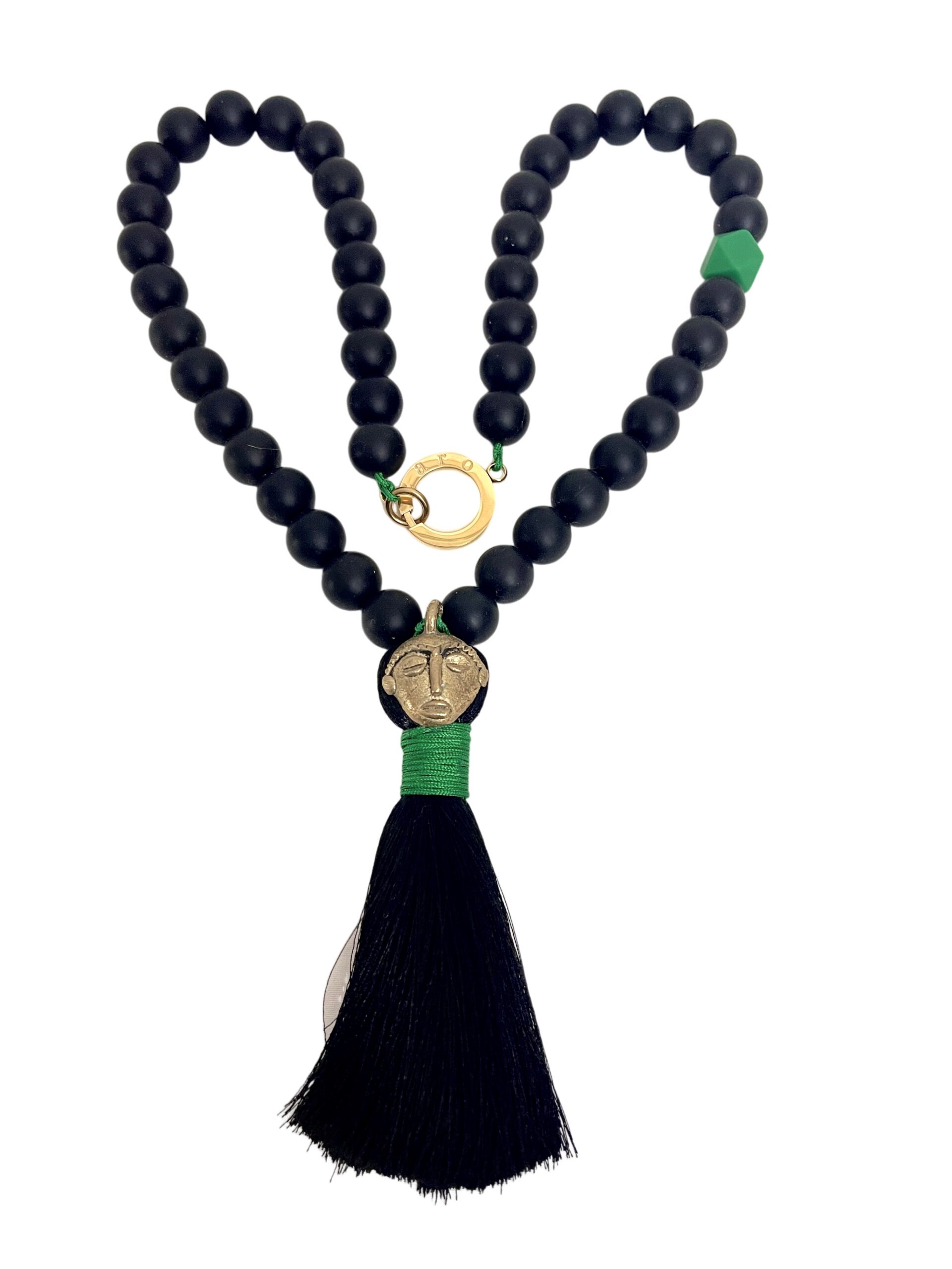 Handcrafted tribal-style necklace featuring matte black silicone beads strung together with green thread, accented by a single green geometric bead and a gold circular clasp. The centerpiece includes a brass face-shaped pendant wrapped with green thread, from which hangs a long black silk tassel.