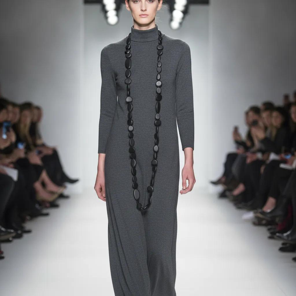 Model walking down a runway wearing a long, charcoal-gray turtleneck dress paired with an extra-long matte black bead necklace. The necklace features irregular oval beads that extend nearly to the knees, creating a minimalist yet dramatic silhouette.
