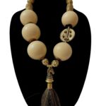 A dramatic statement necklace displaying a horsetail pendant suspended by extra-large ivory color beads, coconut shells, and brass beads.   The necklace is made with smaller beads on the back of the neck for comfort.  The necklace measures 28IN/23OZ.  It is an impressive look in a color that is easy to wear.