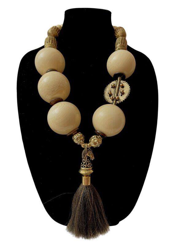 A dramatic statement necklace displaying a horsetail pendant suspended by extra-large ivory color beads, coconut shells, and brass beads.   The necklace is made with smaller beads on the back of the neck for comfort.  The necklace measures 28IN/23OZ.  It is an impressive look in a color that is easy to wear.