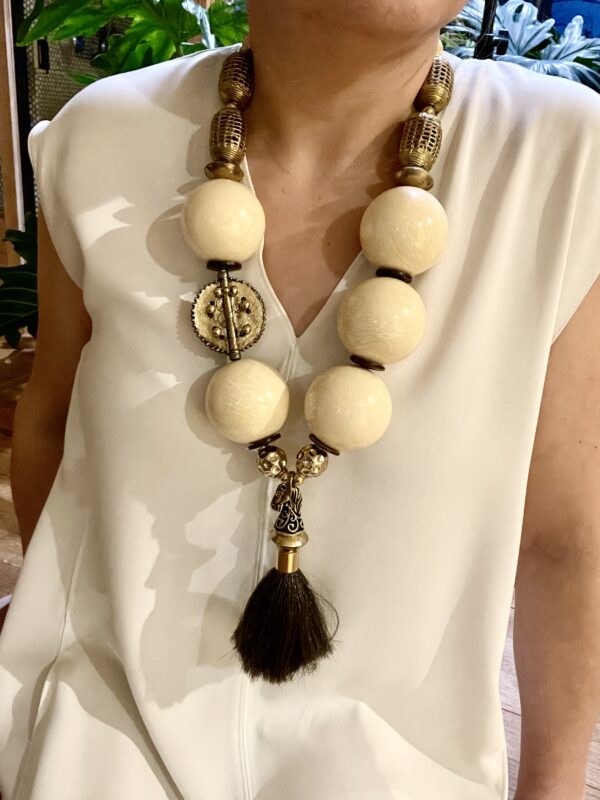 A dramatic statement necklace displaying a horsetail pendant suspended by extra-large ivory color beads, coconut shells, and brass beads.   The necklace is made with smaller beads on the back of the neck for comfort.  The necklace measures 28IN/23OZ.  It is an impressive look in a color that is easy to wear.