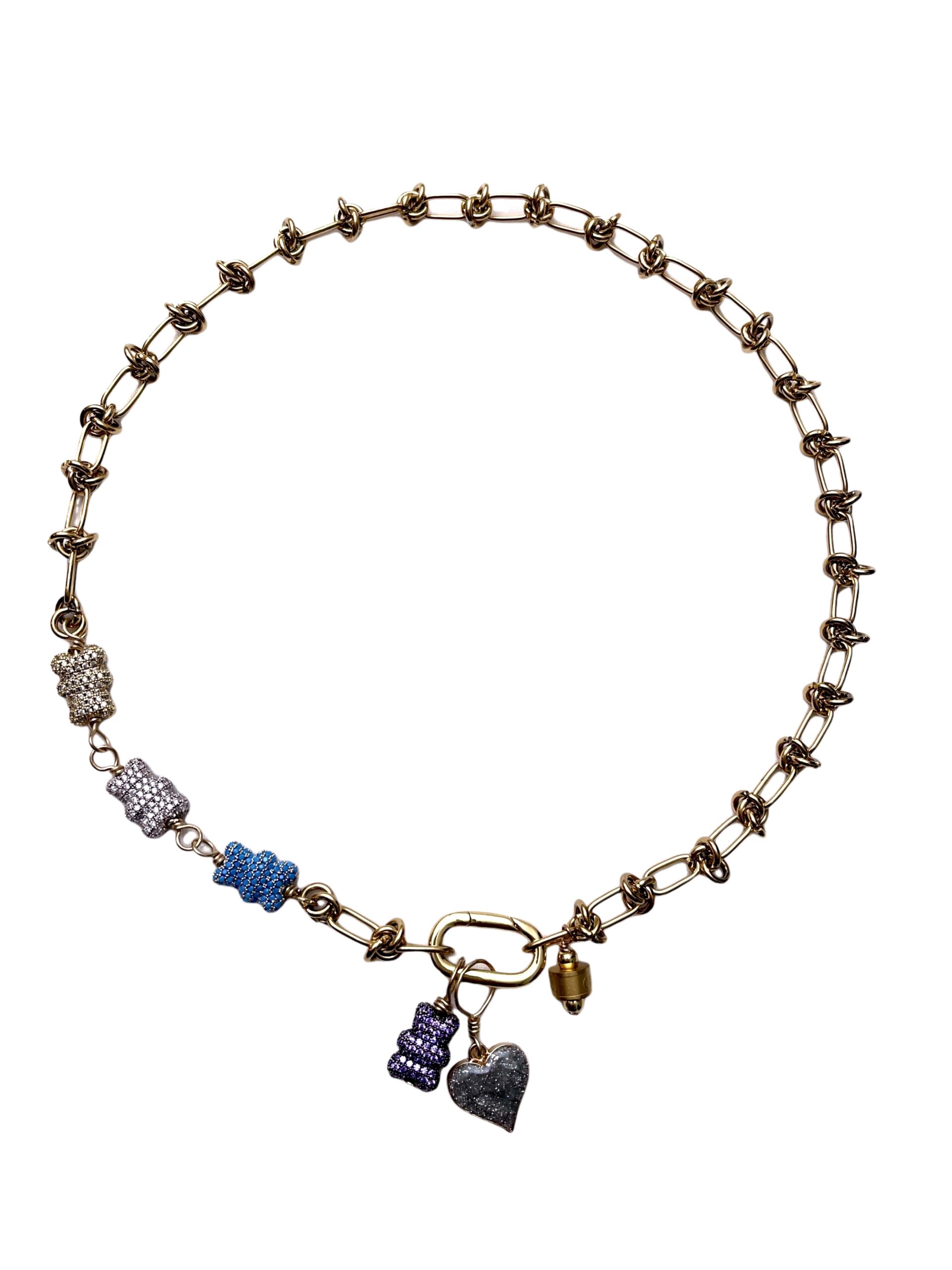 Gold chain necklace featuring interlinked oval and twisted loops with several sparkling charms. The charms include bear-shaped pendants in silver, blue, purple, and gold tones, along with a glittery silver heart and a small cylindrical gold charm near the clasp.