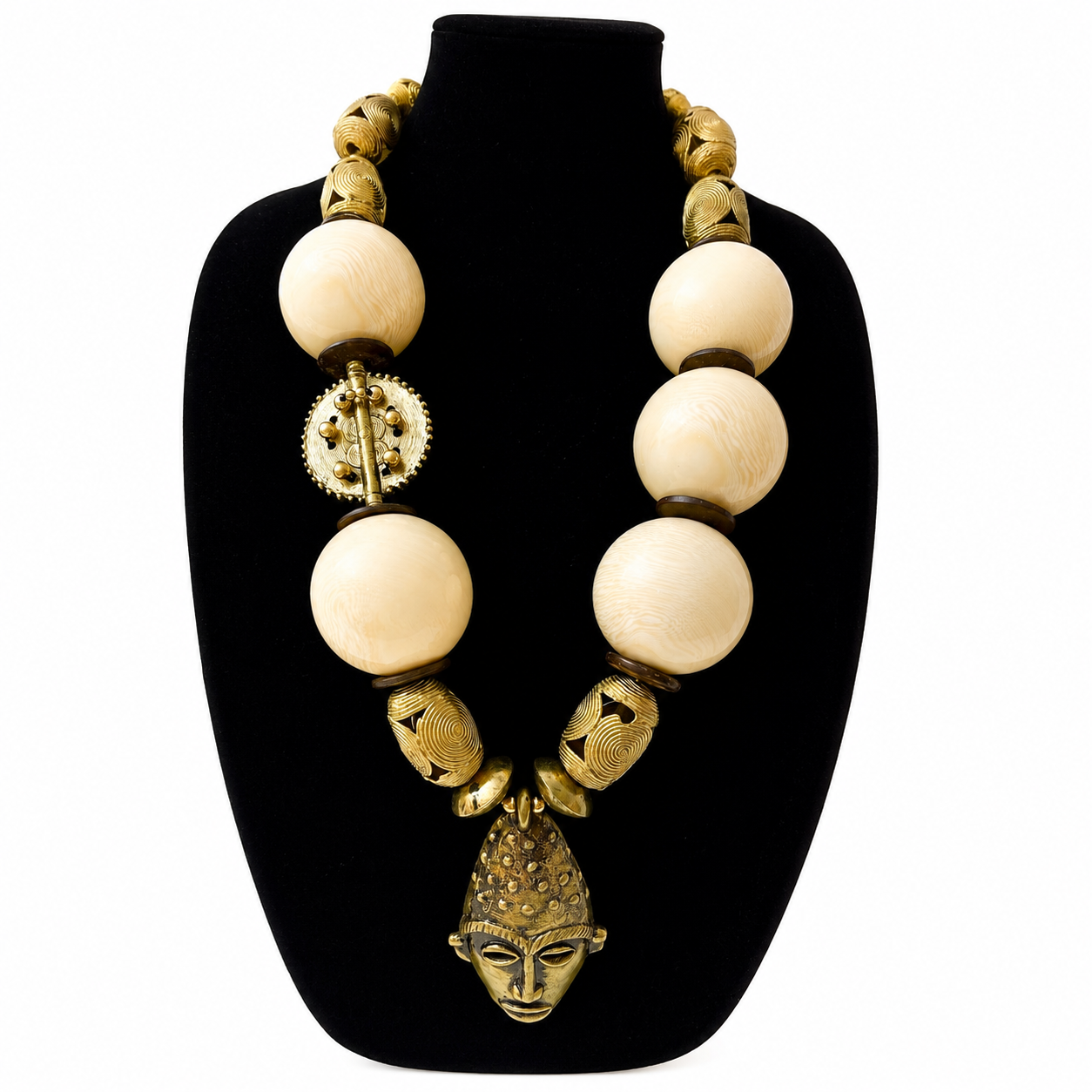 Statement necklace displayed on a black velvet bust, featuring large ivory-colored spherical beads interspersed with ornate brass elements. The centerpiece is a sculpted brass mask pendant with intricate detailing and a textured headdress, evoking traditional African artistry.