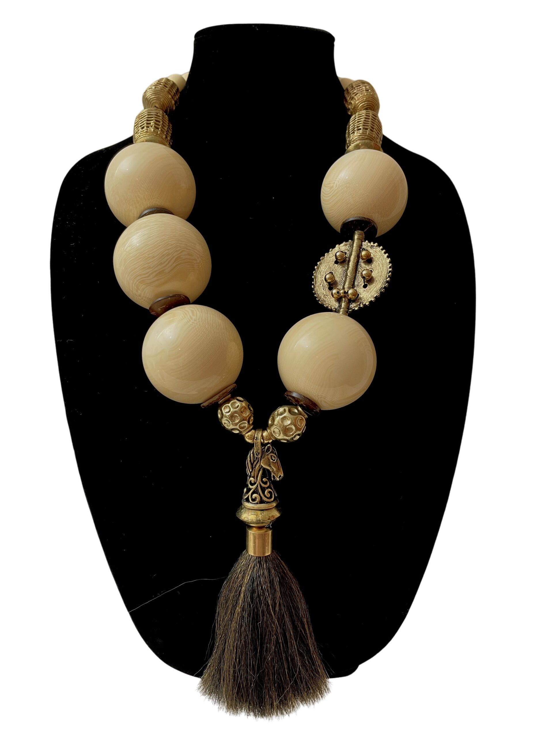 Statement necklace displayed on a black bust, featuring large ivory-colored spherical beads with subtle wood-grain texture. The design includes ornate gold accents, a circular gear-like ornament, and a sculpted horse head pendant with a dark brown tassel.