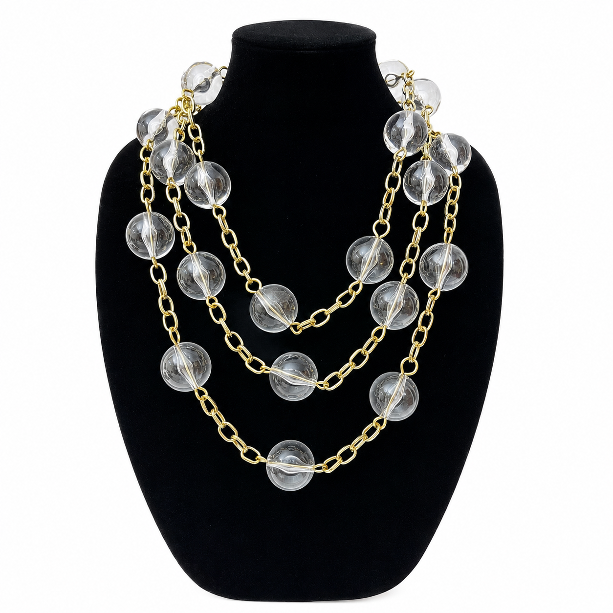 Close-up of the Clear Obsession Necklace displayed on a black velvet bust. The necklace features large transparent spherical beads linked by gold chains, arranged in three cascading layers that create a luminous, sculptural effect.