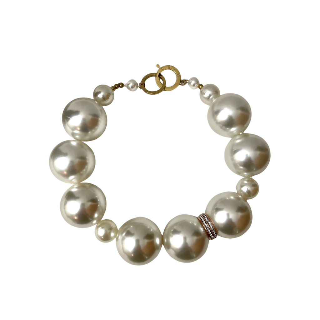 Single-strand pearl necklace featuring large, lustrous white pearls arranged in a graduated pattern. One pearl near the center is accented with a slim, gold-toned ring encrusted with small crystals. The necklace closes with a circular gold clasp engraved with “Caro,” adding a refined touch.