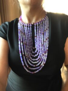 The choker measures 16 IN, and the longest strand of beads measures 30 IN.   The necklace design displays dangling glass beads in shades of purple with brass accent beads—the beads lay in the front of the chest without moving out of place. 