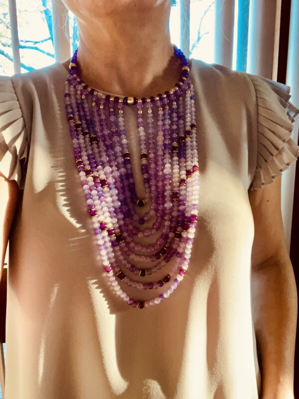 The choker measures 16 IN, and the longest strand of beads measures 30 IN.   The necklace design displays dangling glass beads in shades of purple with brass accent beads—the beads lay in the front of the chest without moving out of place. 