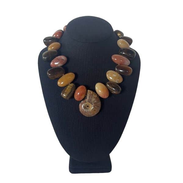 A rich blend of natural texture and earthy elegance, this necklace showcases the warm, grounded character of genuine Tiger Eye paired with the soft, muted tones of Aventurine and the ancient beauty of a single large Ammonite fossil. Measuring 18 inches in length and weighing 11 ounces, it offers a substantial, sculptural presence while resting comfortably along the neckline. The keyword Rusty appears naturally in the palette, reflected in the warm browns, golden flashes, and mineral striations that define Tiger Eye’s signature look. Tiger Eye is known for its chatoyant shimmer—bands of light that move across the stone like liquid gold. Historically, it has been associated with stability, courage, and protection, qualities often linked to its deep, Rusty glow and grounding energy. Aventurine adds a complementary earth‑tone softness, creating a harmonious balance between sheen and matte texture. At the center, the Ammonite fossil introduces a dramatic focal point, its spiral form symbolizing evolution, resilience, and the passage of time. Each element is arranged to highlight the necklace’s natural materials, giving it an organic yet refined aesthetic. The combination of Tiger Eye’s Rusty warmth, Aventurine’s subtle hues, and the fossil’s ancient geometry makes this piece a striking statement for collectors and lovers of earth‑born design.