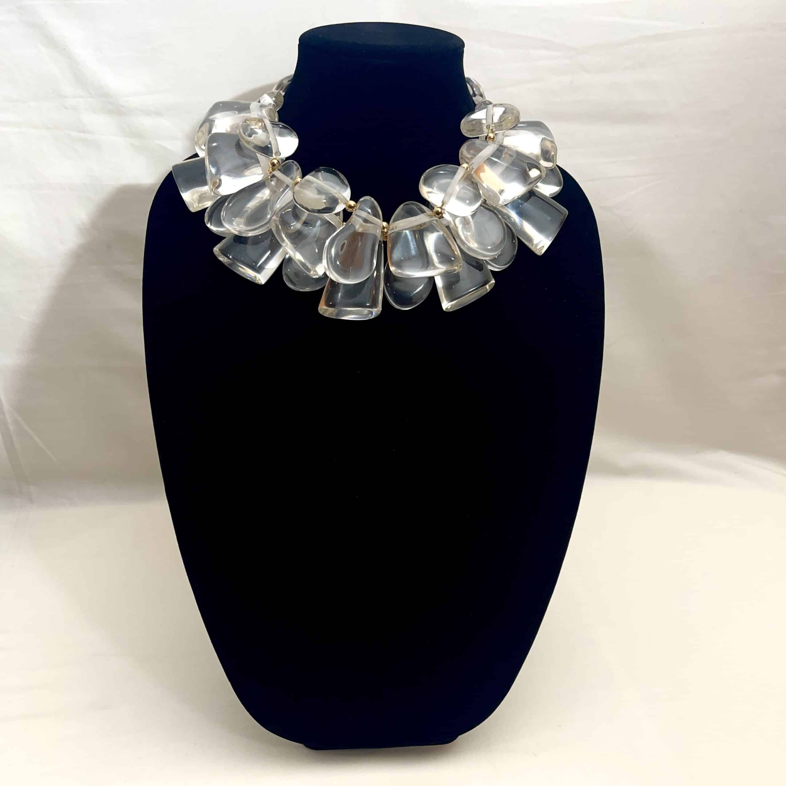 Clear Acrylic Cluster Necklace with Gold Accents Make a bold impression with this sculptural statement necklace, designed to evoke the crystalline beauty of ice.  Featuring a clustered arrangement of three strands of large, clear acrylic beads, this piece catches and reflects light with dazzling clarity.  Each bead is separated by gold-tone spacer accents, adding warmth and contrast to the cool, glass-like shimmer.   The necklace is secured with a large, easy-to-use lobster clasp, ensuring both comfort and durability. Measuring 17 inches in length with an adjustable extension up to 22 inches, it offers versatility for different necklines and styling preferences. At 16 ounces, it has a substantial feel—like wearing a piece of modern art. Whether you're dressing for a gallery opening, a winter soirée, or want to elevate your everyday look, this necklace will turn heads.  Its unique design mimics the elegance of ice formations, while the gold accents lend a touch of luxury.  This is more than jewelry—it’s a wearable sculpture, crafted to impress and inspire.  With its ice-like clarity and bold silhouette, it’s the perfect fusion of art and fashion.