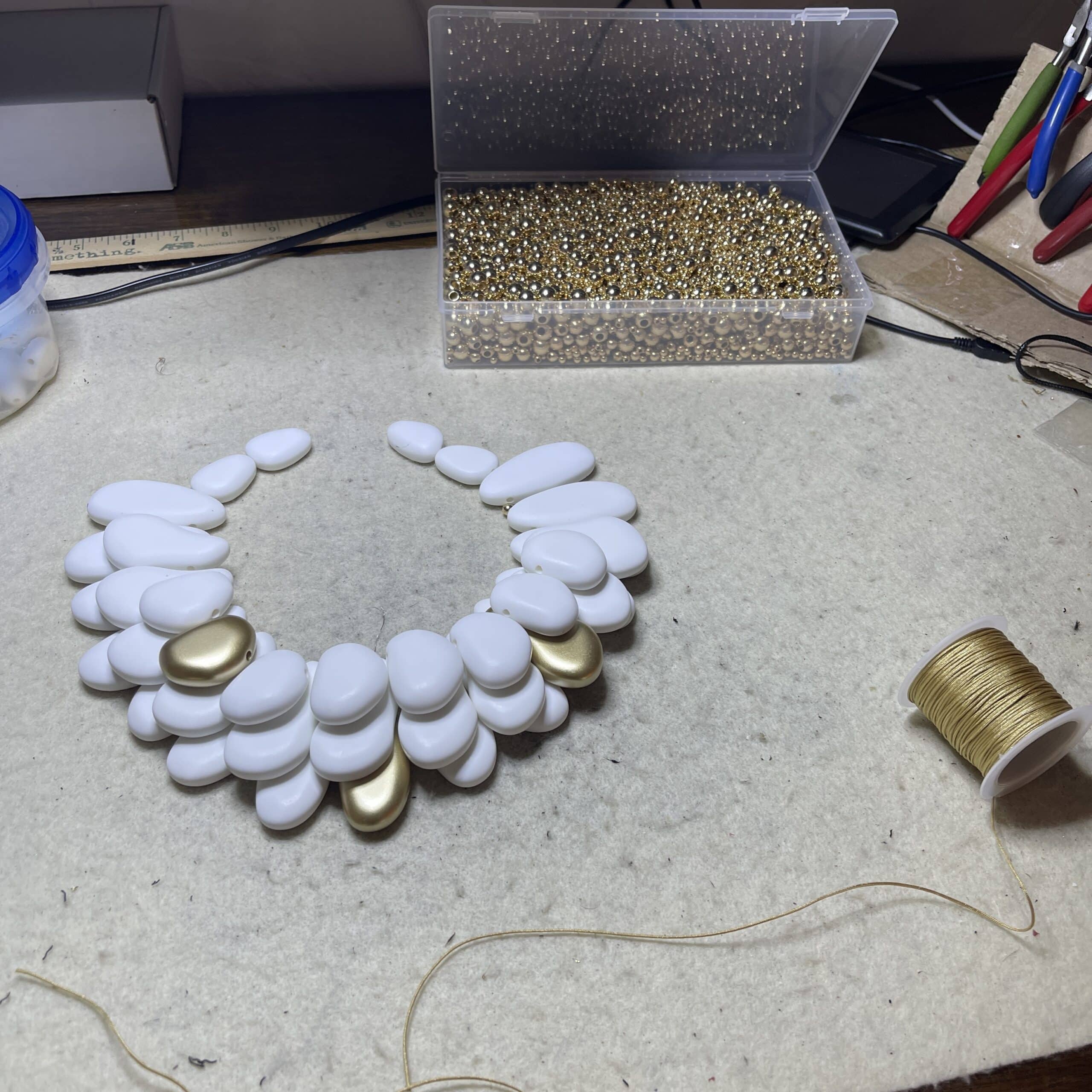 Winter White & Gold Acrylic Bead Necklace Command attention with this regal statement necklace, a sculptural blend of winter white and gleaming gold accents. Crafted with multilayered strands of acrylic beads, the design evokes natural pebbles—smooth, matte, and artfully arranged.  Interspersed among the white beads are gold-toned spacers, adding warmth and contrast to the cool palette. This necklace is more than an accessory—it’s a wearable art piece. The layered construction creates depth and movement, while the circular clasp ensures a secure and polished finish. Measuring 16 inches with an extension up to 21 inches, it adapts effortlessly to different necklines and occasions.  At 14 ounces, it has a bold, confident presence without compromising comfort. Whether paired with a crisp blouse or a dramatic evening dress, this necklace transitions seamlessly from day to night. Its regal tones and organic shapes make it a standout piece for gallery events, holiday gatherings, or everyday elegance. Celebrate your individuality with a design that’s both modern and timeless. The regal interplay of white and gold speaks to strength, grace, and artistic flair—perfect for those who love jewelry that makes a statement.