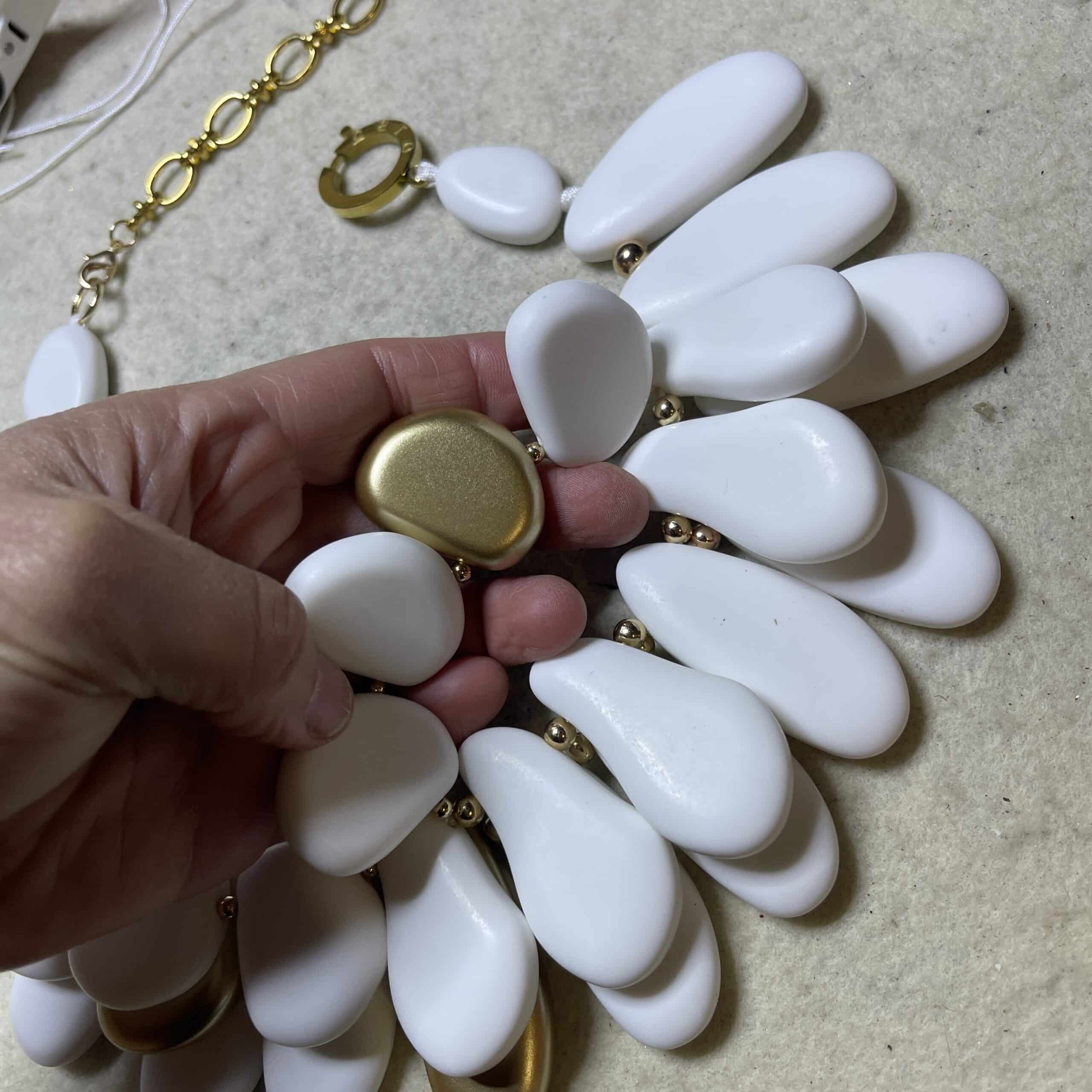 Winter White & Gold Acrylic Bead Necklace Command attention with this regal statement necklace, a sculptural blend of winter white and gleaming gold accents. Crafted with multilayered strands of acrylic beads, the design evokes natural pebbles—smooth, matte, and artfully arranged.  Interspersed among the white beads are gold-toned spacers, adding warmth and contrast to the cool palette. This necklace is more than an accessory—it’s a wearable art piece. The layered construction creates depth and movement, while the circular clasp ensures a secure and polished finish. Measuring 16 inches with an extension up to 21 inches, it adapts effortlessly to different necklines and occasions.  At 14 ounces, it has a bold, confident presence without compromising comfort. Whether paired with a crisp blouse or a dramatic evening dress, this necklace transitions seamlessly from day to night. Its regal tones and organic shapes make it a standout piece for gallery events, holiday gatherings, or everyday elegance. Celebrate your individuality with a design that’s both modern and timeless. The regal interplay of white and gold speaks to strength, grace, and artistic flair—perfect for those who love jewelry that makes a statement.