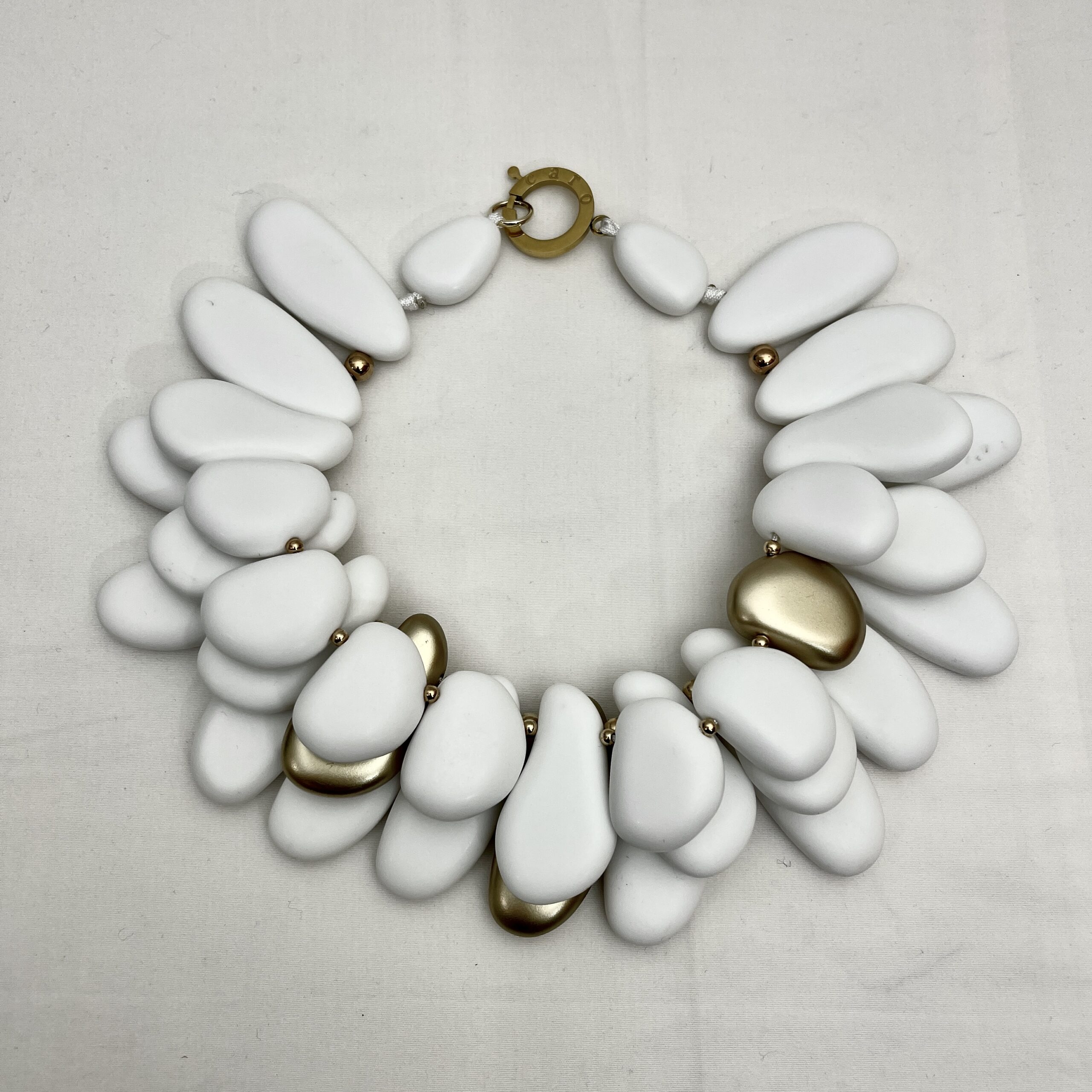 Winter White & Gold Acrylic Bead Necklace Command attention with this regal statement necklace, a sculptural blend of winter white and gleaming gold accents. Crafted with multilayered strands of acrylic beads, the design evokes natural pebbles—smooth, matte, and artfully arranged.  Interspersed among the white beads are gold-toned spacers, adding warmth and contrast to the cool palette. This necklace is more than an accessory—it’s a wearable art piece. The layered construction creates depth and movement, while the circular clasp ensures a secure and polished finish. Measuring 16 inches with an extension up to 21 inches, it adapts effortlessly to different necklines and occasions.  At 14 ounces, it has a bold, confident presence without compromising comfort. Whether paired with a crisp blouse or a dramatic evening dress, this necklace transitions seamlessly from day to night. Its regal tones and organic shapes make it a standout piece for gallery events, holiday gatherings, or everyday elegance. Celebrate your individuality with a design that’s both modern and timeless. The regal interplay of white and gold speaks to strength, grace, and artistic flair—perfect for those who love jewelry that makes a statement.