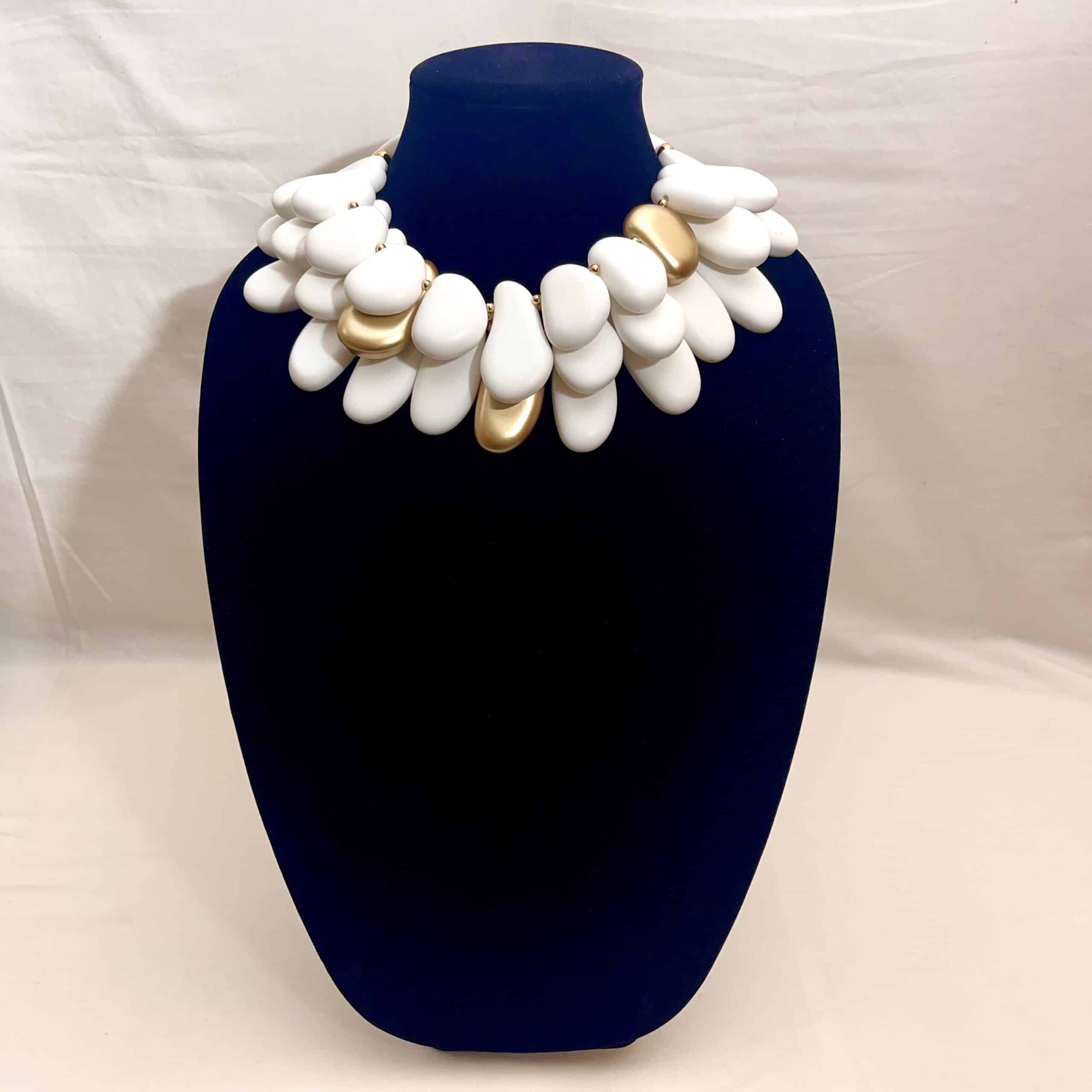 Winter White & Gold Acrylic Bead Necklace Command attention with this regal statement necklace, a sculptural blend of winter white and gleaming gold accents. Crafted with multilayered strands of acrylic beads, the design evokes natural pebbles—smooth, matte, and artfully arranged.  Interspersed among the white beads are gold-toned spacers, adding warmth and contrast to the cool palette. This necklace is more than an accessory—it’s a wearable art piece. The layered construction creates depth and movement, while the circular clasp ensures a secure and polished finish. Measuring 16 inches with an extension up to 21 inches, it adapts effortlessly to different necklines and occasions.  At 14 ounces, it has a bold, confident presence without compromising comfort. Whether paired with a crisp blouse or a dramatic evening dress, this necklace transitions seamlessly from day to night. Its regal tones and organic shapes make it a standout piece for gallery events, holiday gatherings, or everyday elegance. Celebrate your individuality with a design that’s both modern and timeless. The regal interplay of white and gold speaks to strength, grace, and artistic flair—perfect for those who love jewelry that makes a statement.