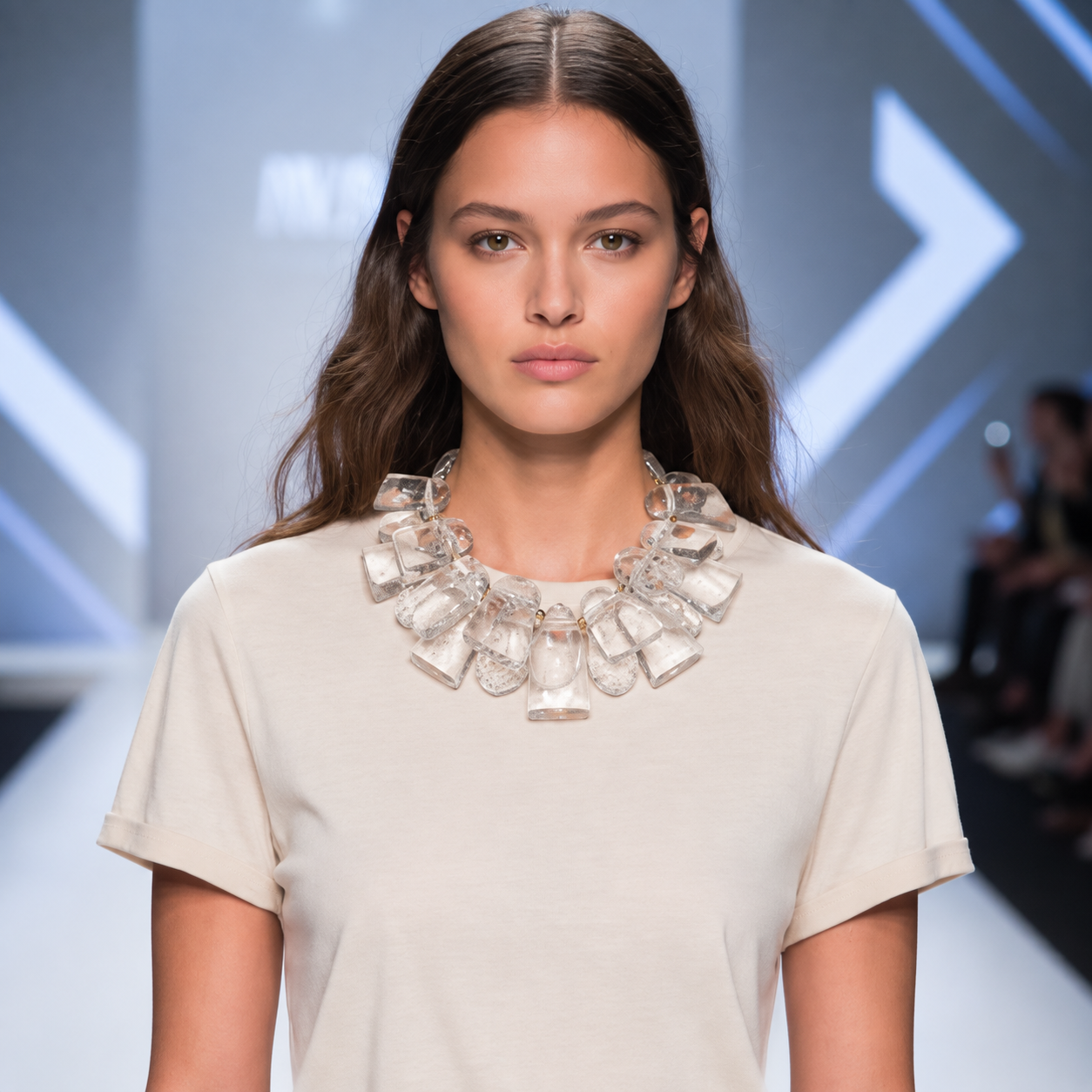 Model walking on a fashion runway wearing a minimalist beige short‑sleeved top and a bold clear acrylic cluster necklace. The necklace features large, irregularly shaped transparent beads that catch the light, creating a sculptural, ice‑like effect. The background shows a blurred runway with geometric patterns and an audience, emphasizing the modern, artistic style of the jewelry.