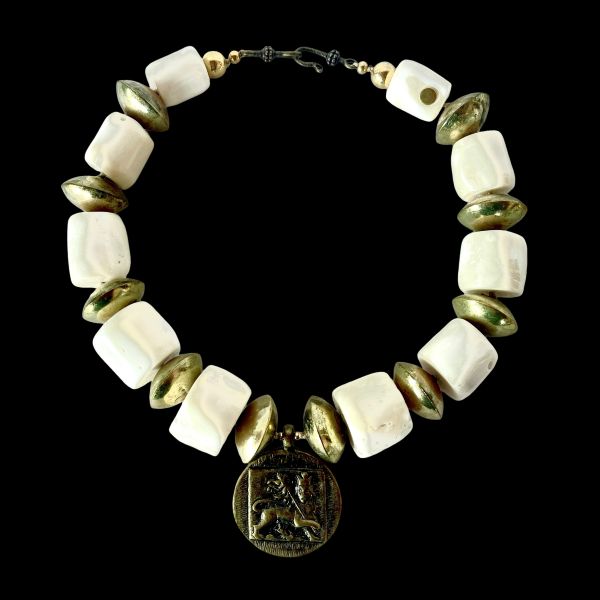 Ostia 0369233 Necklace Strength carved in stone, history cast in brass. This commanding necklace evokes the grandeur of ancient port cities and the power they once held. Crafted with bold white coral beads and substantial brass accents, it channels the elemental contrast of sea and empire—organic purity against forged resilience. At its heart lies a singular medallion: a lion in mid-roar, symbolizing the might and legacy of Ostia during Rome’s golden age. Measuring 17 inches, with an extender reaching 22 inches, this 9-ounce piece sits with presence and poise. The coral beads offer a tactile coolness, while the brass discs catch light like sun on stone. Together, they form a wearable relic—one that speaks of trade routes, mythologies, and the timeless allure of Mediterranean craftsmanship. Whether worn with linen or leather, the Ostia necklace transforms any ensemble into a statement of strength and story. It’s not just jewelry—it’s a tribute to a civilization that revered beauty and power in equal measure. Let the lion guide you. Let the brass gleam like ancient coin. Let Ostia whisper through the ages, three times over, in the language of adornment. Details: Length: 17–22 inches Weight: 9 oz Materials: White coral, brass, lion medallion Style: Heritage statement piece