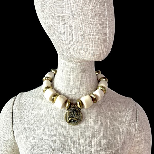 Ostia 0369233 Necklace Strength carved in stone, history cast in brass. This commanding necklace evokes the grandeur of ancient port cities and the power they once held. Crafted with bold white coral beads and substantial brass accents, it channels the elemental contrast of sea and empire—organic purity against forged resilience. At its heart lies a singular medallion: a lion in mid-roar, symbolizing the might and legacy of Ostia during Rome’s golden age. Measuring 17 inches, with an extender reaching 22 inches, this 9-ounce piece sits with presence and poise. The coral beads offer a tactile coolness, while the brass discs catch light like sun on stone. Together, they form a wearable relic—one that speaks of trade routes, mythologies, and the timeless allure of Mediterranean craftsmanship. Whether worn with linen or leather, the Ostia necklace transforms any ensemble into a statement of strength and story. It’s not just jewelry—it’s a tribute to a civilization that revered beauty and power in equal measure. Let the lion guide you. Let the brass gleam like ancient coin. Let Ostia whisper through the ages, three times over, in the language of adornment. Details: Length: 17–22 inches Weight: 9 oz Materials: White coral, brass, lion medallion Style: Heritage statement piece