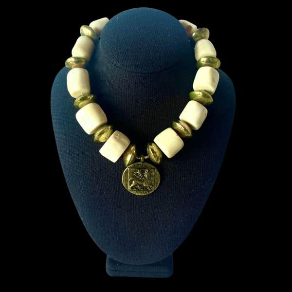 Ostia 0369233 Necklace Strength carved in stone, history cast in brass. This commanding necklace evokes the grandeur of ancient port cities and the power they once held. Crafted with bold white coral beads and substantial brass accents, it channels the elemental contrast of sea and empire—organic purity against forged resilience. At its heart lies a singular medallion: a lion in mid-roar, symbolizing the might and legacy of Ostia during Rome’s golden age. Measuring 17 inches, with an extender reaching 22 inches, this 9-ounce piece sits with presence and poise. The coral beads offer a tactile coolness, while the brass discs catch light like sun on stone. Together, they form a wearable relic—one that speaks of trade routes, mythologies, and the timeless allure of Mediterranean craftsmanship. Whether worn with linen or leather, the Ostia necklace transforms any ensemble into a statement of strength and story. It’s not just jewelry—it’s a tribute to a civilization that revered beauty and power in equal measure. Let the lion guide you. Let the brass gleam like an ancient coin. Let Ostia whisper through the ages, three times over, in the language of adornment. Details: Length: 17–22 inches Weight: 9 oz Materials: White coral, brass, lion medallion Style: Heritage statement piece
