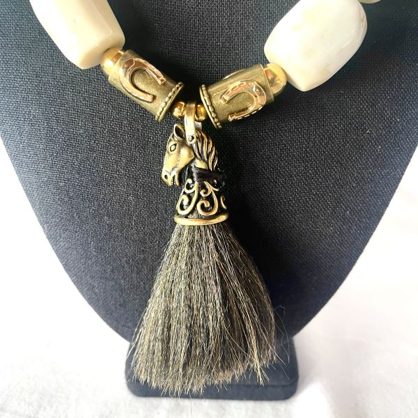 Horseshoe Necklace Where wild grace meets crafted power. This striking necklace blends equestrian spirit with artisanal elegance. Made of large, white natural coral beads and bold brass accents, it carries the weight of tradition and the freedom of open terrain. At 10 ounces and 19 inches long—extending to 24 inches—it drapes with confident presence, each element chosen for symbolism and strength. The clasp features horseshoe-adorned beads, a nod to protection and fortune. These same beads anchor the centerpiece: a finely sculpted brass horse head, detailed with reverence and crowned by a tassel of dark horsehair. It’s a tactile tribute to movement, loyalty, and untamed beauty. This piece isn’t just jewelry—it’s a talisman. A wearable homage to the bond between rider and steed, between earth and spirit. Whether paired with denim or silk, the Horseshoe necklace commands attention and tells a story of resilience, grace, and grounded elegance.  As the brass patina with time, it makes a better contrast with the white coral. Let the coral cool your skin. Let the brass catch the light. Let the Horseshoe guide your stride—three times over, in the language of adornment. Details: Length: 19–24 inches Weight: 10 oz Materials: Natural white coral, brass, horsehair tassel, horseshoe beads Style: Equestrian heritage statement piece