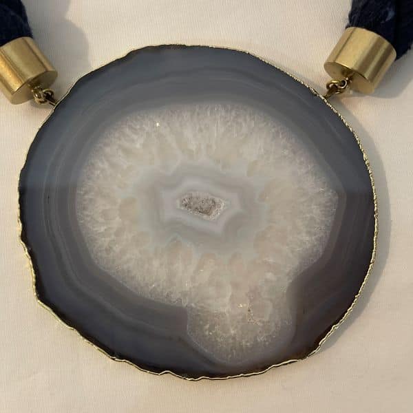 Elliptical Necklace Galactic mystery, grounded in stone. This captivating necklace centers around a large sliced agate pendant, its mineral veins swirling in hypnotic patterns that evoke the illusion of a distant galaxy. The stone’s natural formation—layered in translucent whites, smoky grays, and crystalline shimmer—draws the eye inward, like a portal to something cosmic and ancient. It’s a piece that speaks to both the vastness of space and the intimacy of earthbound craft. Measuring 21 inches and weighing 9 ounces, the necklace balances boldness with elegance. The pendant is suspended from a thick, twisted navy rope, anchored by brass caps and a circular clasp that adds a touch of refinement. The textures—soft rope, cool stone, gleaming metal—create a tactile experience as rich as its visual impact. This is not just jewelry—it’s a meditation on form, time, and the elliptical paths we trace through life. Whether worn with minimalist silhouettes or layered over dramatic fabrics, the Elliptical necklace becomes a focal point, a conversation starter, a quiet echo of the stars. Let the veins guide you. Let the stone speak. Let Elliptical shimmer—three times over, in the language of design. Details: Length: 21 inches Weight: 9 oz Materials: Sliced agate, navy rope, gold-toned clasp Style: Cosmic statement piece with natural elegance