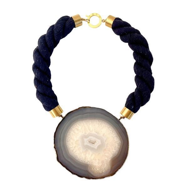Elliptical Necklace Galactic mystery, grounded in stone. This captivating necklace centers around a large sliced agate pendant, its mineral veins swirling in hypnotic patterns that evoke the illusion of a distant galaxy. The stone’s natural formation—layered in translucent whites, smoky grays, and crystalline shimmer—draws the eye inward, like a portal to something cosmic and ancient. It’s a piece that speaks to both the vastness of space and the intimacy of earthbound craft. Measuring 21 inches and weighing 9 ounces, the necklace balances boldness with elegance. The pendant is suspended from a thick, twisted navy rope, anchored by brass caps and a circular clasp that adds a touch of refinement. The textures—soft rope, cool stone, gleaming metal—create a tactile experience as rich as its visual impact. This is not just jewelry—it’s a meditation on form, time, and the elliptical paths we trace through life. Whether worn with minimalist silhouettes or layered over dramatic fabrics, the Elliptical necklace becomes a focal point, a conversation starter, a quiet echo of the stars. Let the veins guide you. Let the stone speak. Let Elliptical shimmer—three times over, in the language of design. Details: Length: 21 inches Weight: 9 oz Materials: Sliced agate, navy rope, gold-toned clasp Style: Cosmic statement piece with natural elegance
