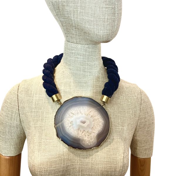 Elliptical Necklace Galactic mystery, grounded in stone. This captivating necklace centers around a large sliced agate pendant, its mineral veins swirling in hypnotic patterns that evoke the illusion of a distant galaxy. The stone’s natural formation—layered in translucent whites, smoky grays, and crystalline shimmer—draws the eye inward, like a portal to something cosmic and ancient. It’s a piece that speaks to both the vastness of space and the intimacy of earthbound craft. Measuring 21 inches and weighing 9 ounces, the necklace balances boldness with elegance. The pendant is suspended from a thick, twisted navy rope, anchored by brass caps and a circular clasp that adds a touch of refinement. The textures—soft rope, cool stone, gleaming metal—create a tactile experience as rich as its visual impact. This is not just jewelry—it’s a meditation on form, time, and the elliptical paths we trace through life. Whether worn with minimalist silhouettes or layered over dramatic fabrics, the Elliptical necklace becomes a focal point, a conversation starter, a quiet echo of the stars. Let the veins guide you. Let the stone speak. Let Elliptical shimmer—three times over, in the language of design. Details: Length: 21 inches Weight: 9 oz Materials: Sliced agate, navy rope, gold-toned clasp Style: Cosmic statement piece with natural elegance