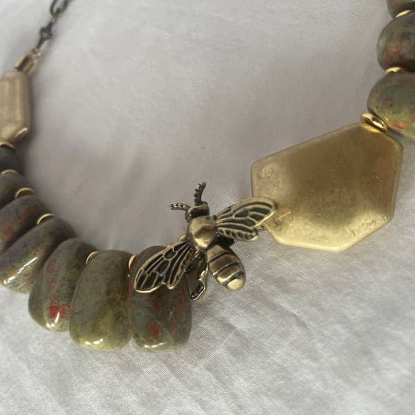 Honey 0369238 is crafted with reverence for nature and history. This 18-inch necklace is a tribute to the enduring beauty of the Appalachian landscape. Each polished Unakite bead—an earthy fusion of green epidote and pink feldspar—is carefully selected for its rich, mottled texture. The red pigments within the stone are thoughtfully echoed by the red cord threading the necklace, creating a warm, harmonious palette. Unakite was first discovered in the Great Smoky Mountains, a region steeped in Cherokee heritage and geological wonder, straddling western North Carolina and eastern Tennessee. Golden accent beads flank a central bee ornament, symbolizing industriousness and the sweetness of life. In ancient cultures, bees were revered as messengers between worlds and guardians of sacred knowledge. This piece channels that legacy, offering a wearable talisman of transformation and abundance. Weighing 12 ounces, the necklace carries a satisfying heft, grounding the wearer in both style and symbolism. Whether worn as a statement piece or a subtle nod to nature’s artistry, it evokes the golden warmth of Honey—its color, its spirit, and its timeless allure. Let this necklace be your daily drop of Honey, a reminder of the sweetness found in resilience, craftsmanship, and the quiet hum of history.