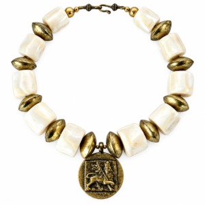 Statement necklace composed of alternating large square white coral beads and oval brass beads. At the center hangs a round brass medallion featuring a raised lion motif within a square frame, symbolizing strength and heritage. The necklace has a hook clasp with small decorative brass spheres.