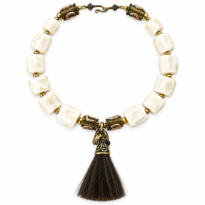 Equestrian-inspired necklace featuring large square white coral beads interspersed with brass spheres and cylindrical beads engraved with horseshoe motifs. At the center hangs a sculpted brass horse head pendant with intricate detailing, crowned by a dark horsehair tassel. The necklace closes with a hook clasp in antique brass finish.