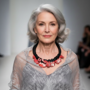 Elegant older woman with silver hair wearing a sheer lace gray top and a striking necklace composed of black cords and red marbled coral pieces. The necklace rests prominently at her collarbone, adding a modern, artistic touch to her refined ensemble.