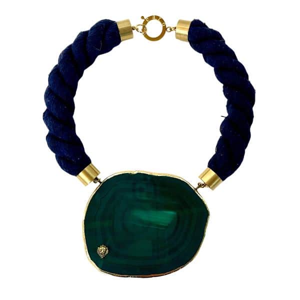 Gaia Necklace Earth’s pulse, captured in stone. This evocative necklace pays homage to the living spirit of the planet. At its center lies a polished green gemstone—its deep hue and swirling bands formed by mineral forces over millennia. The result is a natural phenomenon that personifies the Earth itself, a visual echo of forests, rivers, and ancient terrain. Framed in gold-toned metal and suspended from a thick, twisted navy cord, the piece balances raw beauty with refined craftsmanship. Measuring 21 inches and weighing 8 ounces, the Gaia necklace rests with quiet strength. The brass end caps and circular clasp add a touch of warmth, while the bold cord anchors the design in modern elegance. It’s a statement piece that speaks of origin, resilience, and reverence. Wear it as a tribute to nature’s artistry. Let the stone remind you of cycles, roots, and renewal. Let Gaia whisper through your wardrobe—three times over, in the language of adornment. Details: Length: 21 inches Weight: 8 oz Materials: Green gemstone, navy cord, gold-toned metal Style: Earth-inspired statement piece with elemental elegance