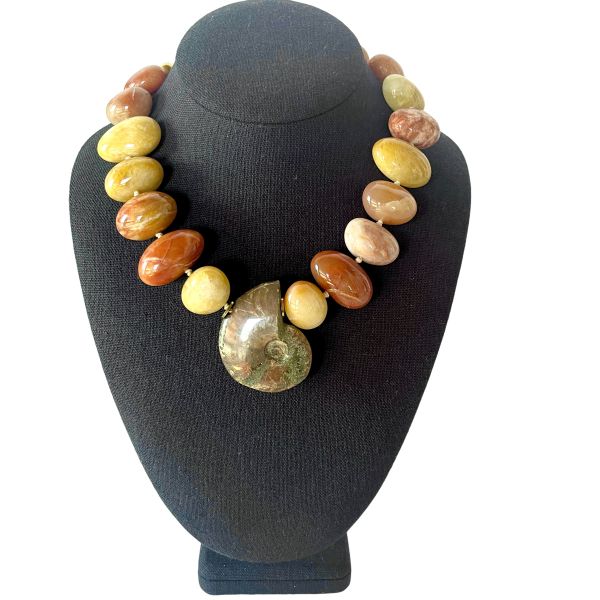 Fossil Necklace Ancient form. Modern radiance. Earth‑born elegance. This striking necklace honors the quiet power of deep time, centered around a beautifully preserved ammonite whose spiraling chambers reveal the mineral artistry of the natural world. Over millions of years, layers of aragonite, calcite, and iron-rich sediments transformed the original shell into a captivating Fossil with intricate patterns and earthy tones that feel both ancient and alive. Highly polished Aventurine beads frame the centerpiece, their warm, luminous hues adding a gentle shimmer that complements the ammonite’s organic geometry. Known for its subtle sparkle and grounding energy, Aventurine brings a refined glow to the design, enhancing its natural sophistication. Measuring 18 inches with an extender reaching 23, the necklace offers a customizable drape, while its 8‑ounce weight provides a satisfying, substantial presence. Each bead and curve reflects the slow, patient work of nature—pressure, heat, and mineral infusion shaping what was once a living creature into a treasured Fossil. The result is a piece that feels both timeless and contemporary, a wearable reminder of Earth’s evolving story. This necklace invites admiration, sparks conversation, and carries the quiet strength of transformation. Let the ammonite’s spiral draw the eye, let the Aventurine glow softly against your skin, and let the ancient beauty of this Fossil accompany you wherever you go.