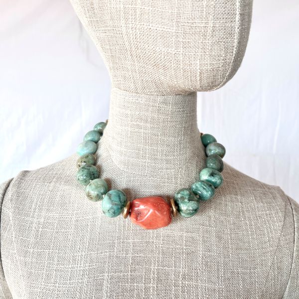 Amazon Necklace Rooted in nature. Radiant in spirit. This necklace is a tribute to the quiet power of simplicity. Crafted with natural amazonite beads in soft green-blue tones, it evokes the lush tranquility of riverbeds and forest canopies. At its heart lies a single coral stone—bold, warm, and unmistakably alive—anchoring the piece with a pulse of vibrant energy. Amazonite, known as the “stone of truth,” is celebrated for its calming influence and its ability to inspire honest expression. Its gentle hues mirror the serenity of the Amazon’s flowing waters, inviting clarity and emotional balance. The necklace’s 18-inch length, extendable to 23 inches, offers graceful drape and versatility, while its 8-ounce weight ensures a substantial yet comfortable presence. Natural coconut spacer beads add an earthy rhythm between the stones, enhancing the organic harmony of the design. A decorative clasp with gold-toned accents completes the look with artisanal finesse. Amazon is more than a name—it’s a feeling, a force, a connection to the wild and the wise. This necklace channels that essence, offering a wearable reminder of strength in softness and truth in simplicity. Let it be your companion in moments of reflection, expression, and quiet confidence.